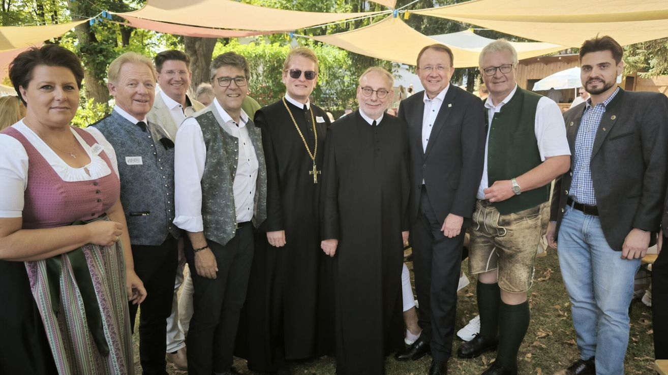 Eine Gruppe von Männern, darunter zwei Priester, stehen zusammen im Freien. Ein Priester trägt eine Kette mit einem Kreuz. Die Männer sind in verschiedenen Kleidungsstücken gekleidet, darunter Anzüge und traditionelle Kleidung. Sie scheinen für ein Foto zu posieren.