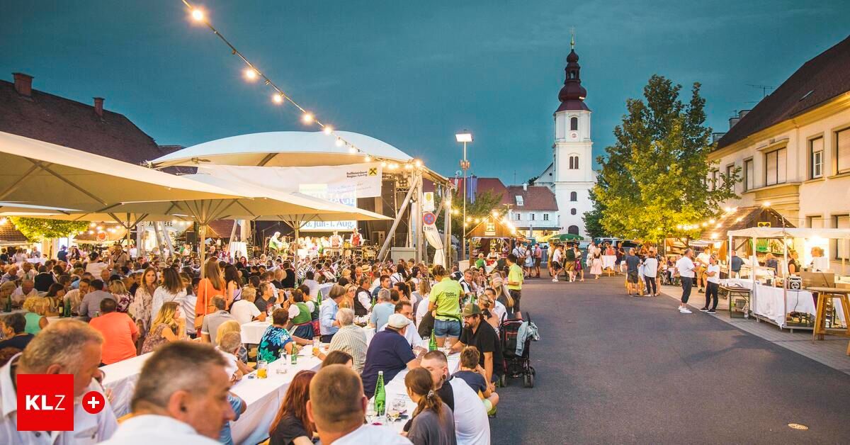 Ein geschäftiges Open-Air-Event mit vielen Menschen an Tischen unter großen Sonnenschirmen, einem Turm im Hintergrund und einem bewölkten Himmel.
