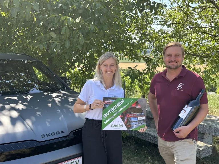 Ein Mann und eine Frau stehen neben einem Skoda Auto, lächeln und halten eine Geschenkbox und eine Flasche Wein. Die Frau hält eine grüne Box mit dem Wort 'Gutschein' darauf.