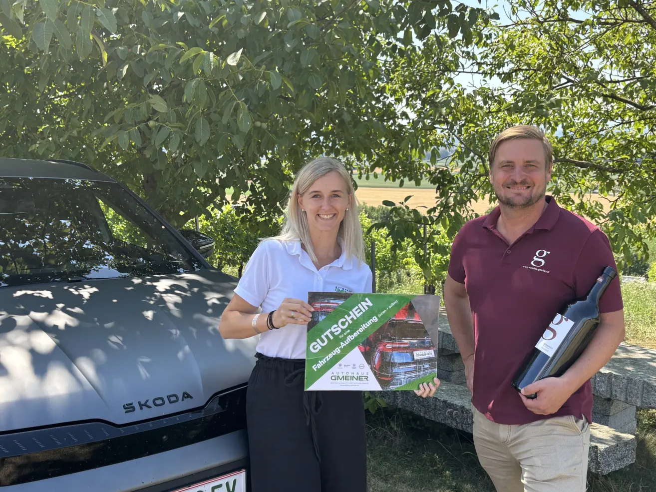 Ein Mann und eine Frau stehen neben einem Skoda Auto, lächeln und halten eine Geschenkbox und eine Flasche Wein. Die Frau hält eine grüne Box mit dem Wort 'Gutschein' darauf.