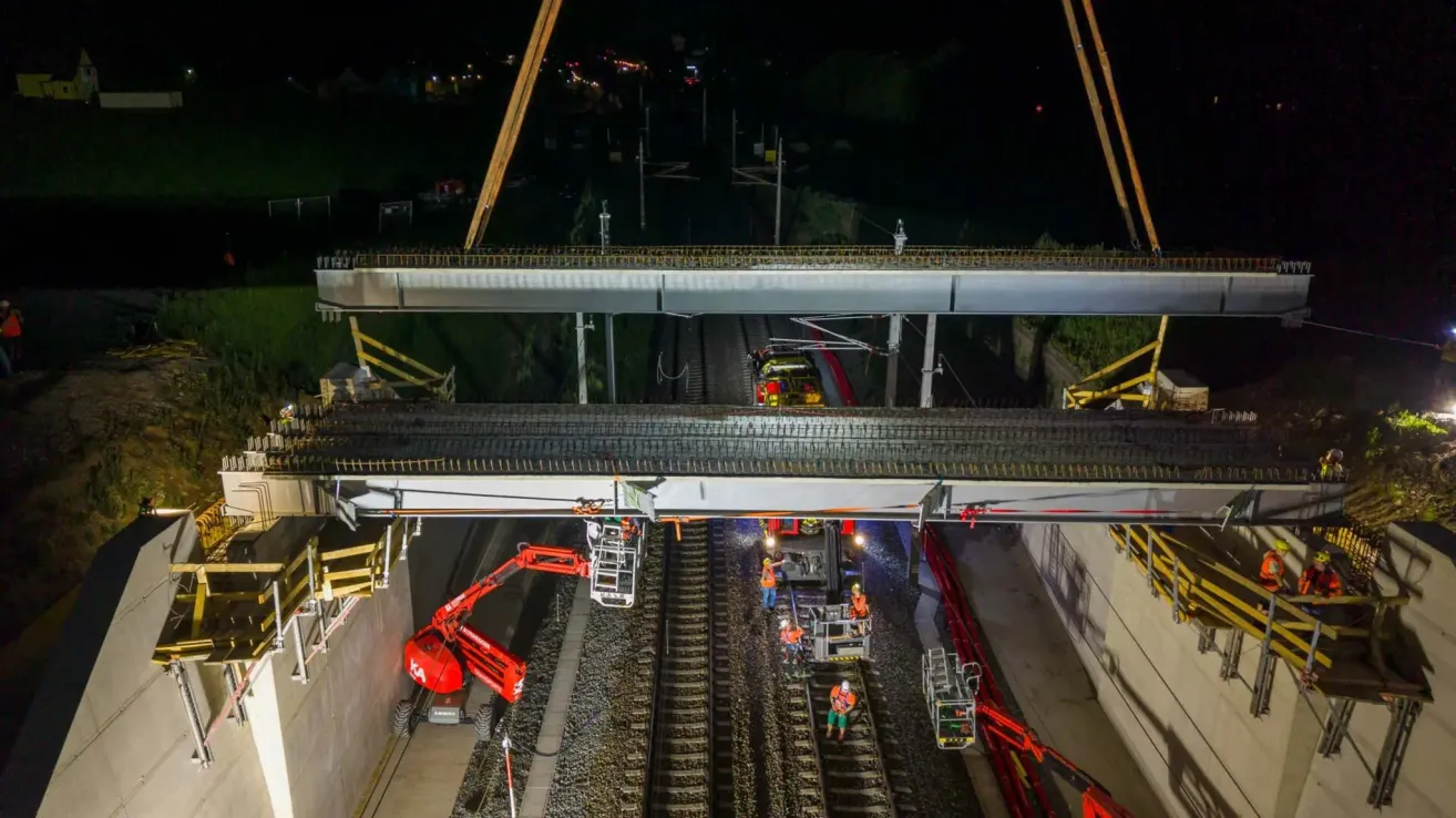 Eine Nachtaufnahme eines Baustellenüberblicks, bei dem Arbeiter eine Brücke über eine Eisenbahnstrecke installieren. Ein roter Kran steht auf der linken Seite, und mehrere Arbeiter sind auf den Gleisen, einige tragen Sicherheitswesten.