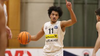 Ein Basketballspieler in einem weißen Trikot mit der Nummer 11, der einen Ball dribbelt und auf etwas zeigt.