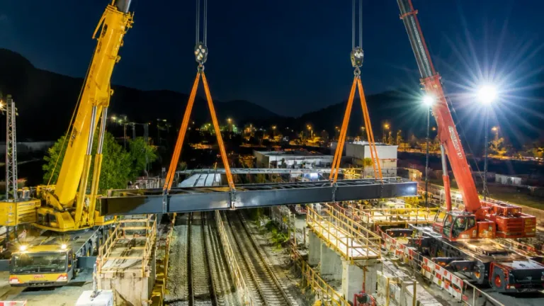 Ein großer Stahlträger wird bei Nacht von zwei Kränen über eine Eisenbahnstrecke gehoben. Im Hintergrund ist eine Stadtsilhouette mit Bergen zu sehen.