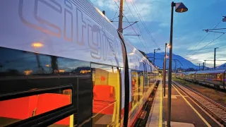 Ein Cityjet-Zug steht an einem Bahnhof, mit roten Sitzen durch die Fenster sichtbar und Bergen im Hintergrund.