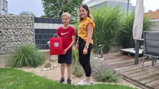 A young boy in a red shirt holds a red bag while standing next to a woman in a yellow top and black pants in a garden.