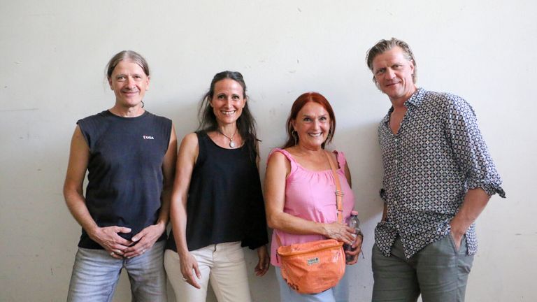 Vier Erwachsene, zwei Männer und zwei Frauen, stehen zusammen vor einer weißen Wand. Der Mann links trägt ein schwarzes Shirt mit Logo, während die Frau rechts eine orange Tasche trägt. Die Frau in der Mitte trägt ein schwarzes Tanktop und weiße Hose.