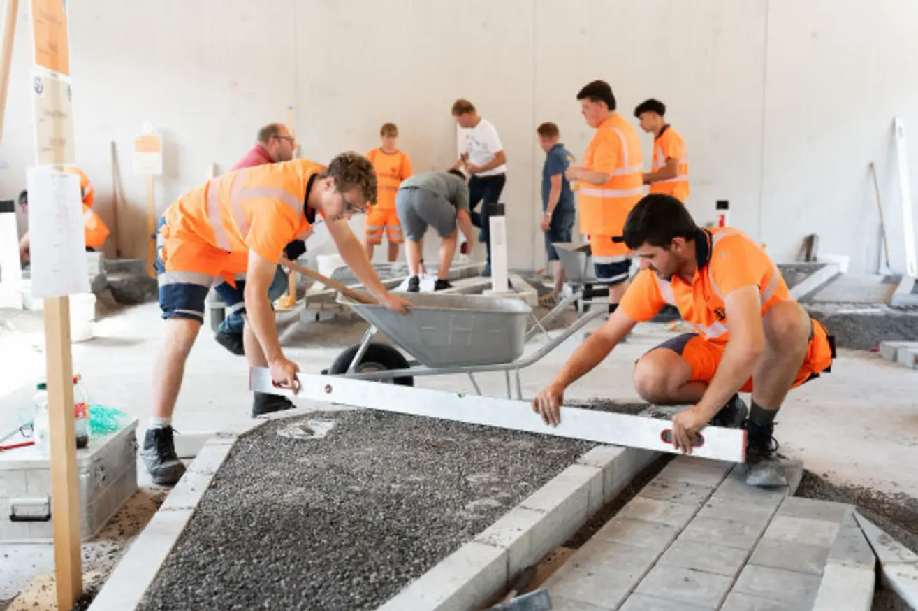 Construction workers in orange uniforms are working on a concrete pathway, one is using a level while another is pushing a wheelbarrow.