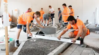 Bauarbeiter in orangefarbenen Uniformen arbeiten an einem Betonweg, einer benutzt ein Lot, während ein anderer einen Schubkarren schiebt.