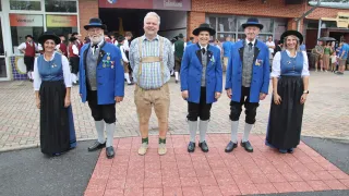 Vier Personen in traditioneller bayerischer Kleidung stehen auf einem roten Ziegelpflaster vor einem Gebäude mit dem Schild 'Thermenlandhalle Kulturhaus'.