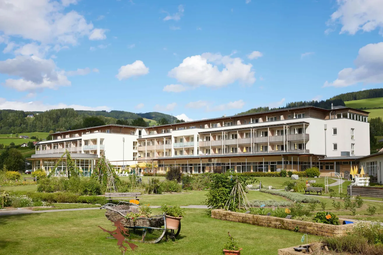 Ein modernes Hotel mit mehreren Stockwerken und Balkonen, umgeben von einem üppigen Garten mit verschiedenen Pflanzen und einem Weg, der zum Eingang führt.