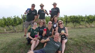 A group of people in green shirts and hats are posing for a photo in a vineyard, holding wine glasses.