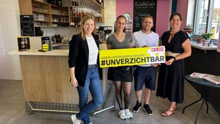 Vier Personen stehen in einem Café und halten ein gelbes Schild mit dem Hashtag #unverzichtbar. Die Frau links trägt einen Blazer, die Frau in der Mitte ein graues Shirt und der Mann rechts ein weißes Shirt. Hinter ihnen steht eine Theke mit einer Tafel.