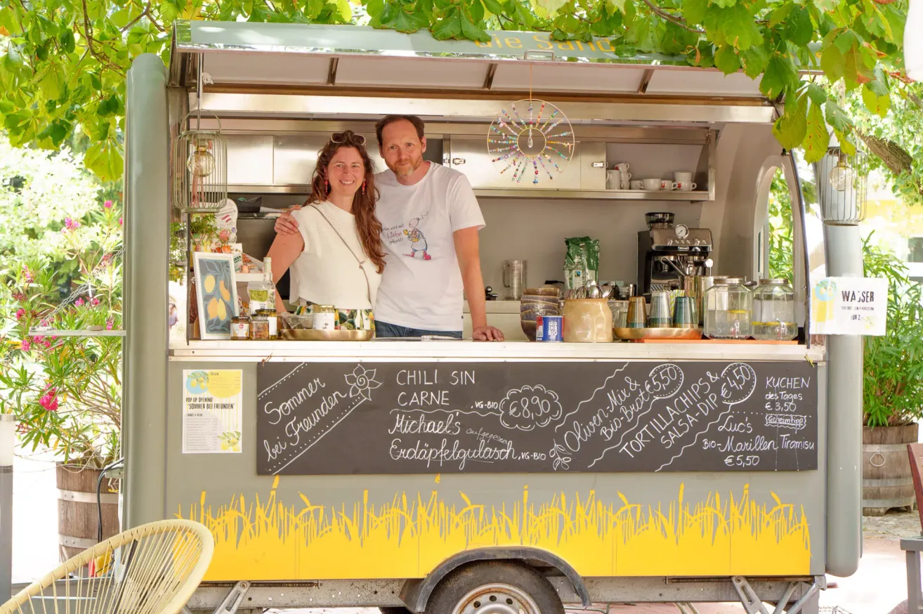 Ein Paar steht vor einem Food-Truck mit einer Kreidetafel-Menü, das verschiedene Gerichte und Preise anbietet.
