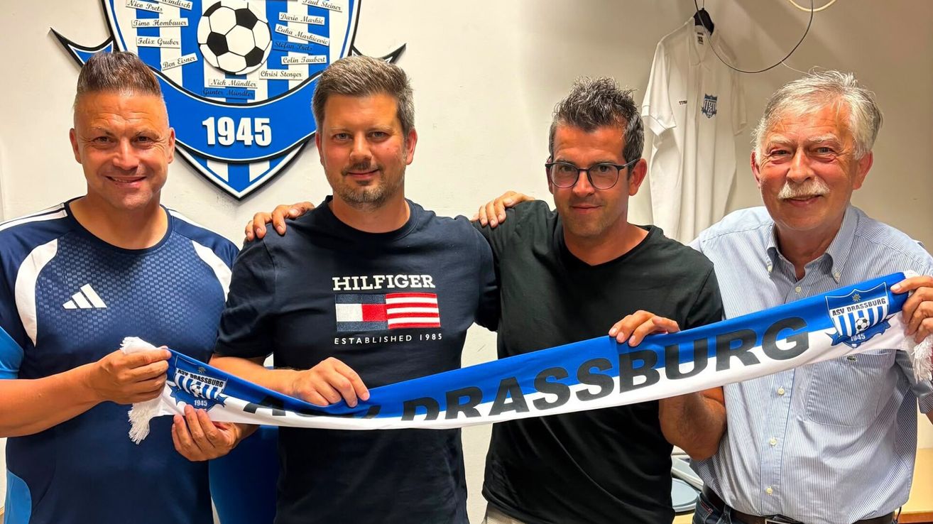 Zwei Männer halten ein Banner mit dem Wort 'DRASSBURG' vor einem Fußballteam-Logo. Der Mann auf der linken Seite trägt ein Hilfiger-Shirt.
