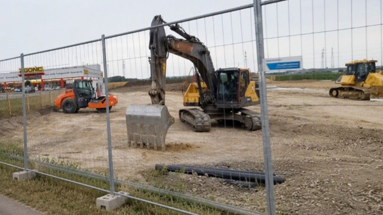 Ein großer gelber und schwarzer Bagger gräbt auf einer Baustelle, umgeben von einem Metallzaun. Ein weiterer Bagger ist in der Nähe geparkt.