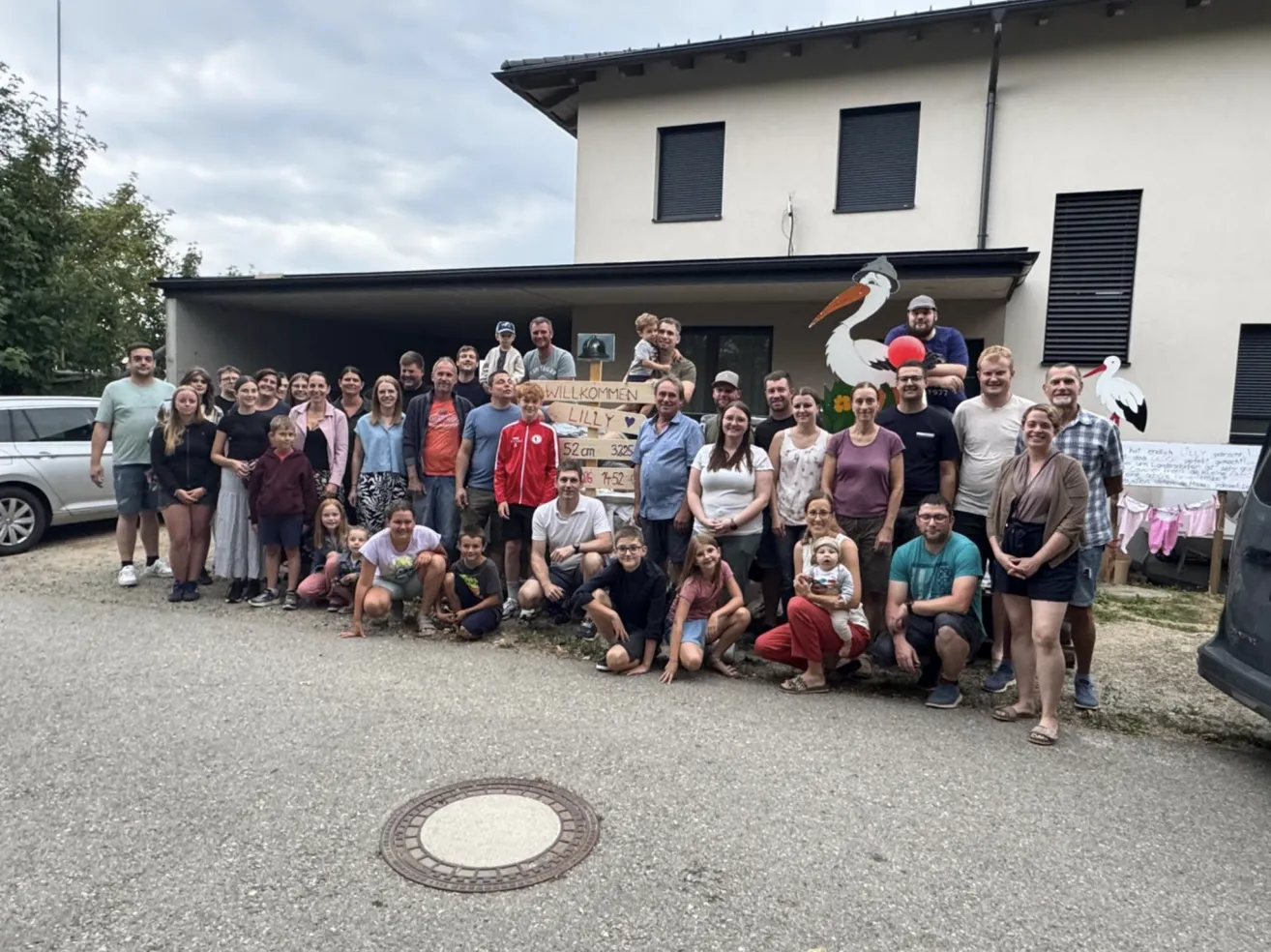 Eine Gruppe von Menschen steht vor einem Haus mit einem Storch-Dekor, einige halten ein Schild mit der Aufschrift 'Willkommen Lilly'. Sie alle lächeln und posieren für ein Foto.