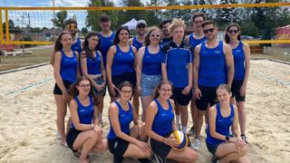 A group of young people in blue jerseys are posing for a photo on the beach, with a volleyball net in the background.