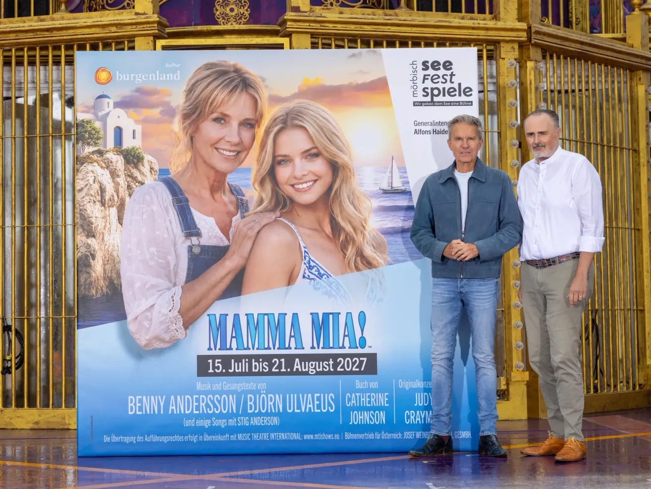 Drei Männer stehen vor einem großen Plakat für das Musical Mamma Mia! Das Poster zeigt zwei Frauen und einen Mann, mit dem Datumsbereich vom 15. Juli bis 21. August 2027.