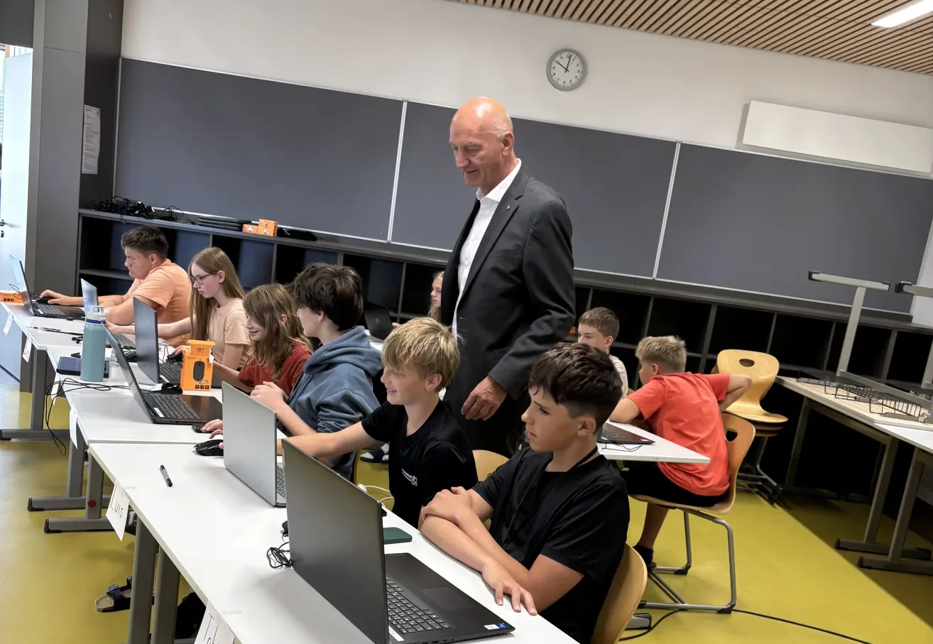 Ein Klassenzimmer mit Schülern, die an Laptops arbeiten, ein Mann im Anzug steht lächelnd hinter ihnen.