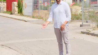 A man in a white shirt and gray pants stands on a street, smiling and gesturing with his hands. Behind him, there are buildings, fences, and a red container.