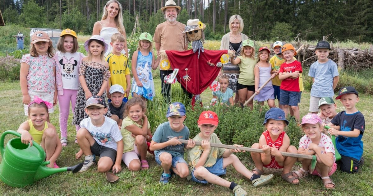 A group of children and adults are posing for a photo in a garden with a scarecrow in the center. The children are wearing hats and holding sticks, while the adults are standing behind them. The garden is surrounded by tall trees.