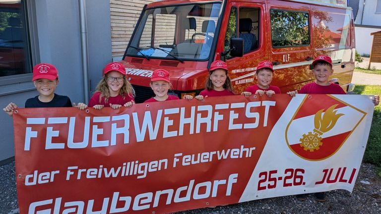 Vier Kinder in roten Mützen und Brillen stehen hinter einem Banner, das Freiwilligen Feuerwehr besagt. Ein roter Feuerwehrwagen ist im Hintergrund sichtbar.
