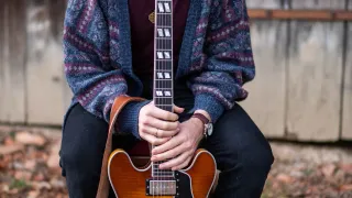 A person is kneeling on the ground, holding a guitar with a strap around their shoulder. They are wearing a knitted sweater and pants.