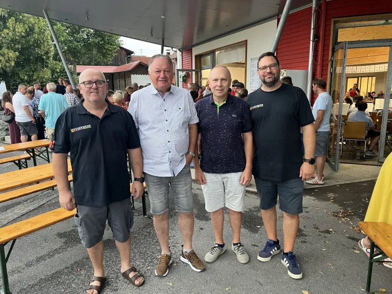Vier Männer stehen vor einem Gebäude, zwei tragen schwarze Shirts mit 'Feuerwehr' darauf. Der Mann in der Mitte trägt ein weißes Hemd und graue Shorts. Der Mann auf der rechten Seite trägt ein schwarzes Hemd und graue Shorts.