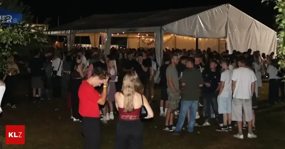 A large crowd of people are gathered at night under a tent with a sign that reads 'MAR'. Some are standing while others are talking. The area is dimly lit.