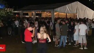 A large crowd of people are gathered at night under a tent with a sign that reads 'MAR'. Some are standing while others are talking. The area is dimly lit.