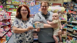 A man and a woman are holding a baby and a check in a store. The man is wearing glasses and a gray shirt, and the woman is wearing a floral shirt. Behind them are shelves with toys and other items.