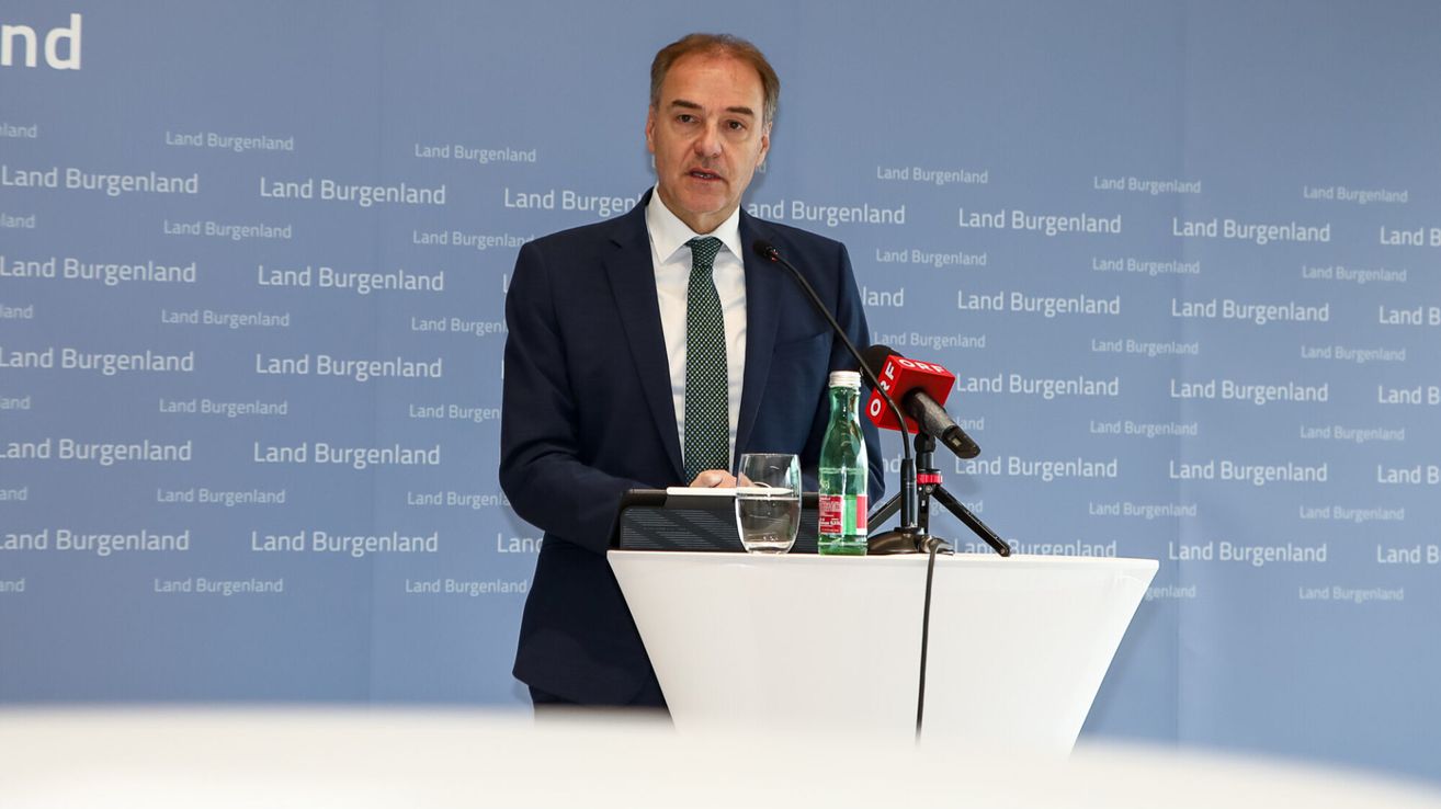 A man in a suit and tie stands at a podium, speaking into a microphone. Behind him is a backdrop with the word 'Burgenland'.