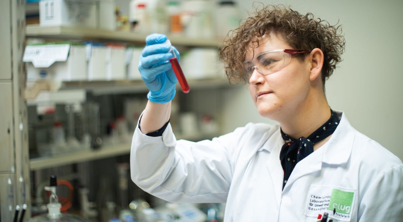 A woman in a lab coat and safety goggles holds a test tube with red liquid inside.