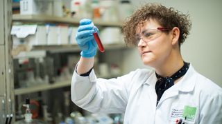 A woman in a lab coat and safety goggles holds a test tube with red liquid inside.