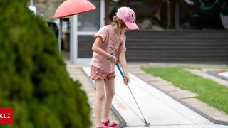 A young girl wearing a pink hat and shirt is playing golf on a sunny day. She is holding a golf club and is about to hit the ball.