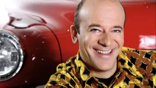 A smiling man with a bald head wearing a yellow and black patterned shirt is standing in front of a red car.