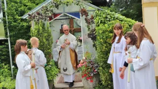 A priest in a white and gold robe holds a silver bell and stands in front of a small chapel, surrounded by children in white robes holding candles.