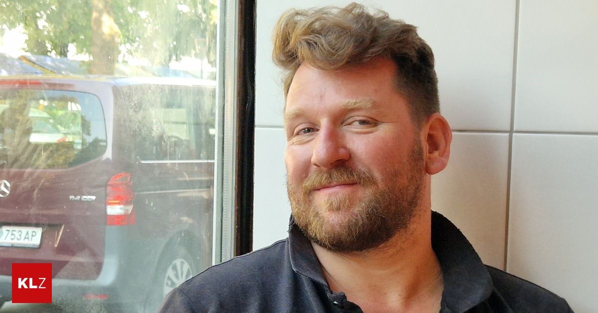 A man with a beard and mustache smiles at the camera, standing in front of a glass window with a car reflection.