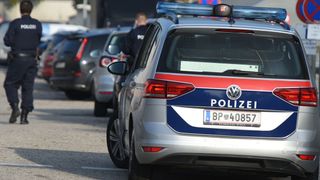 A police car with the license plate BP 40857 is parked in a parking lot with other cars. A police officer is standing behind the car.