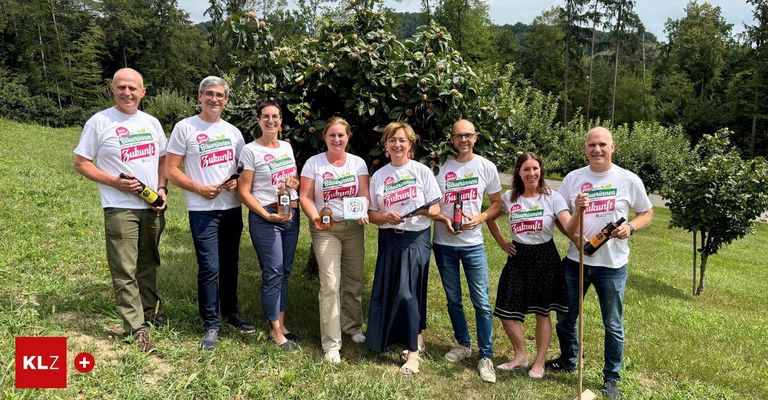 Eine Gruppe von Menschen in passenden weißen T-Shirts mit dem Wort Zukunft stehen auf einer Wiese, lächeln und halten Flaschen. Sie posieren für ein Foto mit einem großen Baum im Hintergrund.