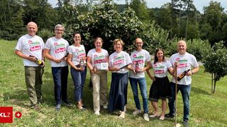 A group of people in matching white t-shirts with the word Zukunft are standing in a field, smiling and holding bottles. They are posing for a photo with a large tree in the background.