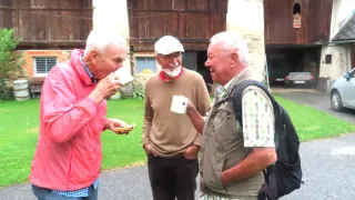 Three elderly men stand outside a rustic building, one drinking from a cup, another holding a cup with a blue design, and the third smiling.