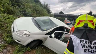 A white car is wrecked on the side of the road with its door open, a firefighter is nearby.