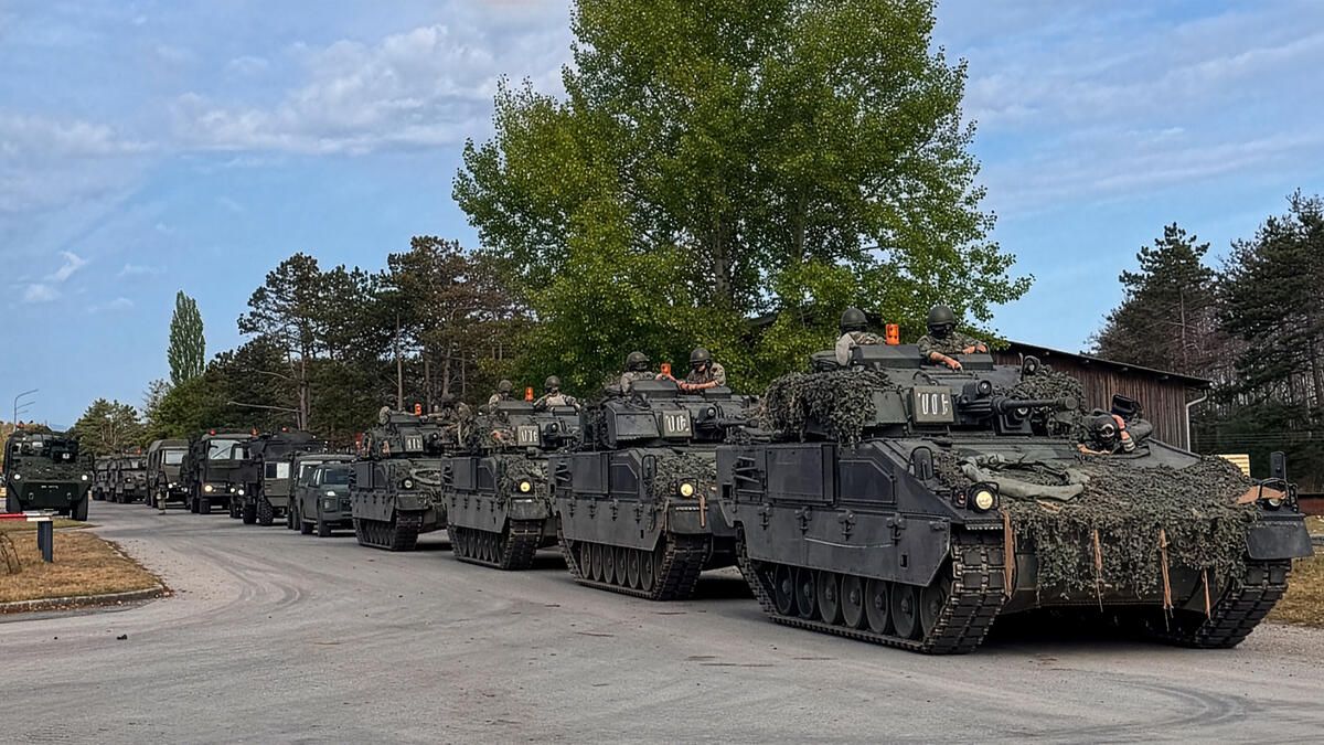 Eine Reihe von militärischen gepanzerten Fahrzeugen mit Soldaten in Helmen steht in einer Reihe auf einer Straße mit Bäumen im Hintergrund.