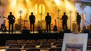 A band performs on stage with a large number 40 in the background. The stage is set with various musical instruments and speakers. The audience is seated in rows of chairs in front of the stage.