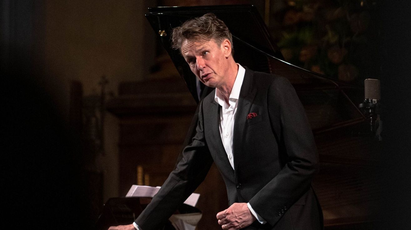 A man in a black suit stands in front of a grand piano, looking to the side with a serious expression.