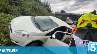 A white car is wrecked on the side of a road with an open door, a person in a reflective vest nearby, and an ambulance in the background.