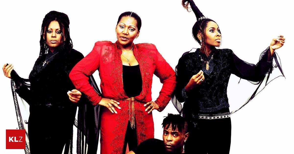 Four individuals are posing for a photograph. The woman in the center is wearing a red dress and has her hands on her hips. The woman on the right is wearing a black dress with a white belt. The man in the front is sitting with his hands on his knees.