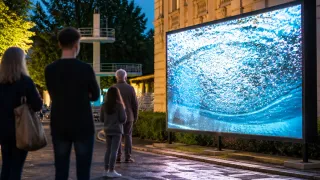 A group of people are standing in front of a large screen showing a water effect, with a building and a diving tower in the background.