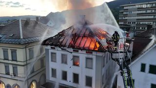 A firefighter stands on a ladder spraying water on a burning building with a damaged roof and windows.